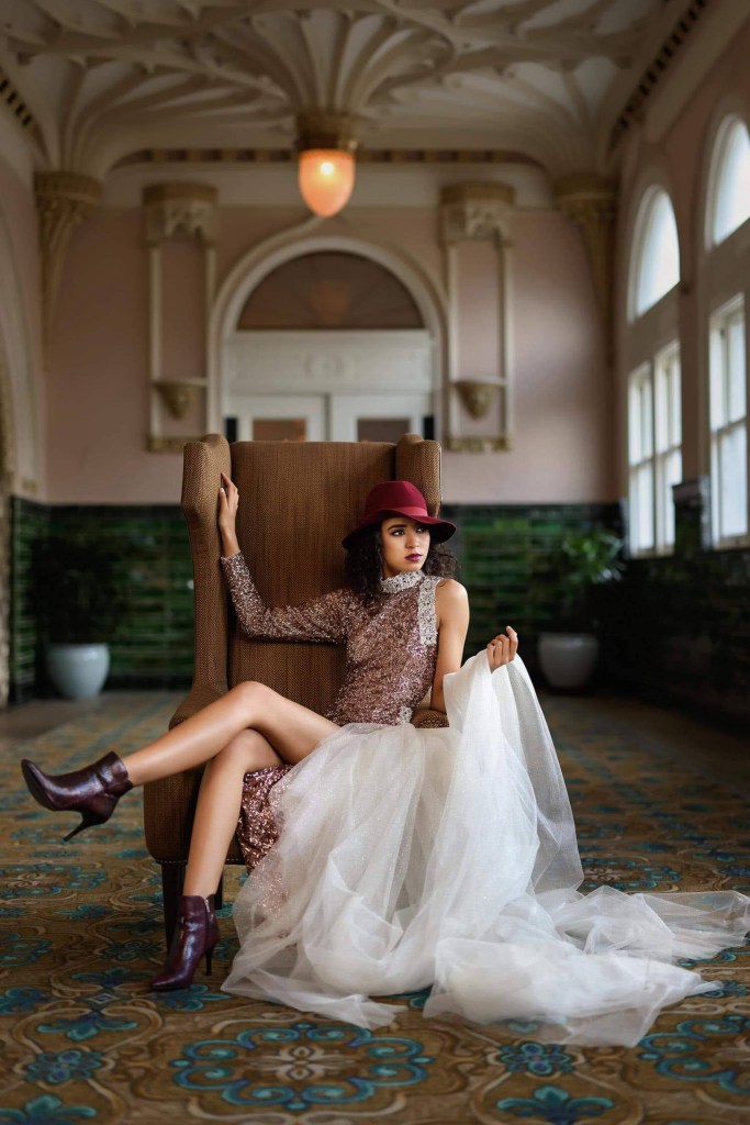 Modella seduta su una poltrona, indossando un abito scintillante e un cappello, con una gonna di tulle bianco che si estende sul pavimento.
